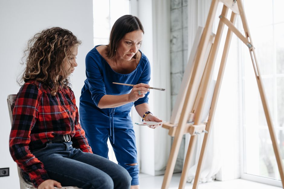 A teacher guides a student during an art class, demonstrating painting techniques on a canvas.